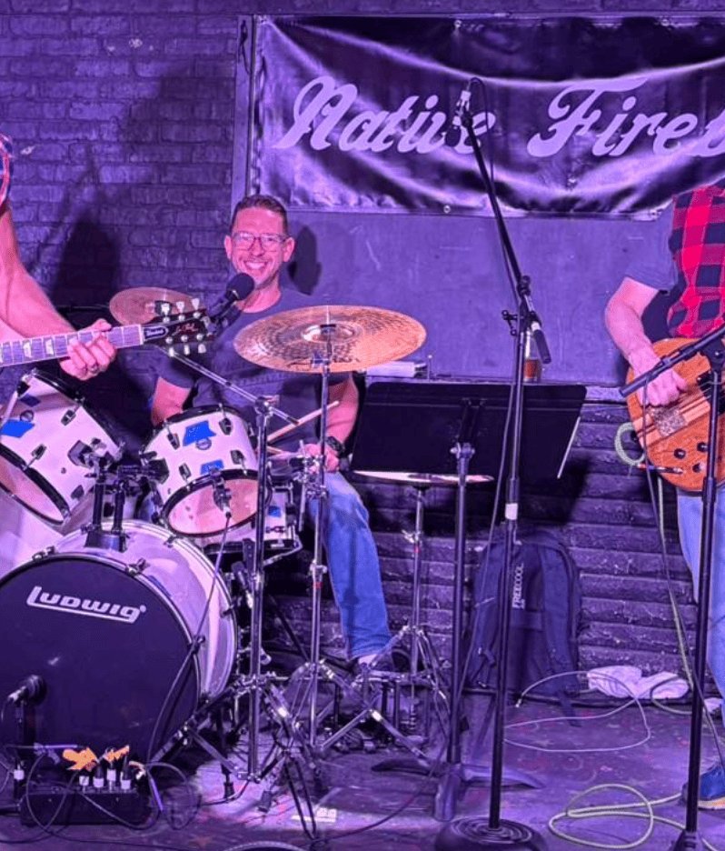 Gabe at a drumkit on a live stage, blue stage lighting, banner behind.